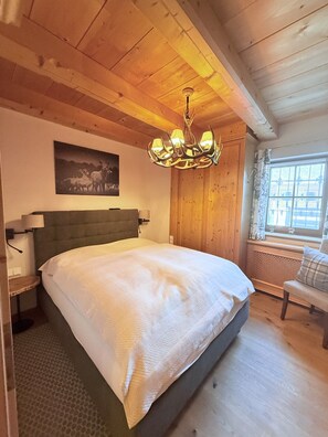 A bedroom with a large bed, wooden ceiling, a chandelier, a window with a view, and a painting on the wall.