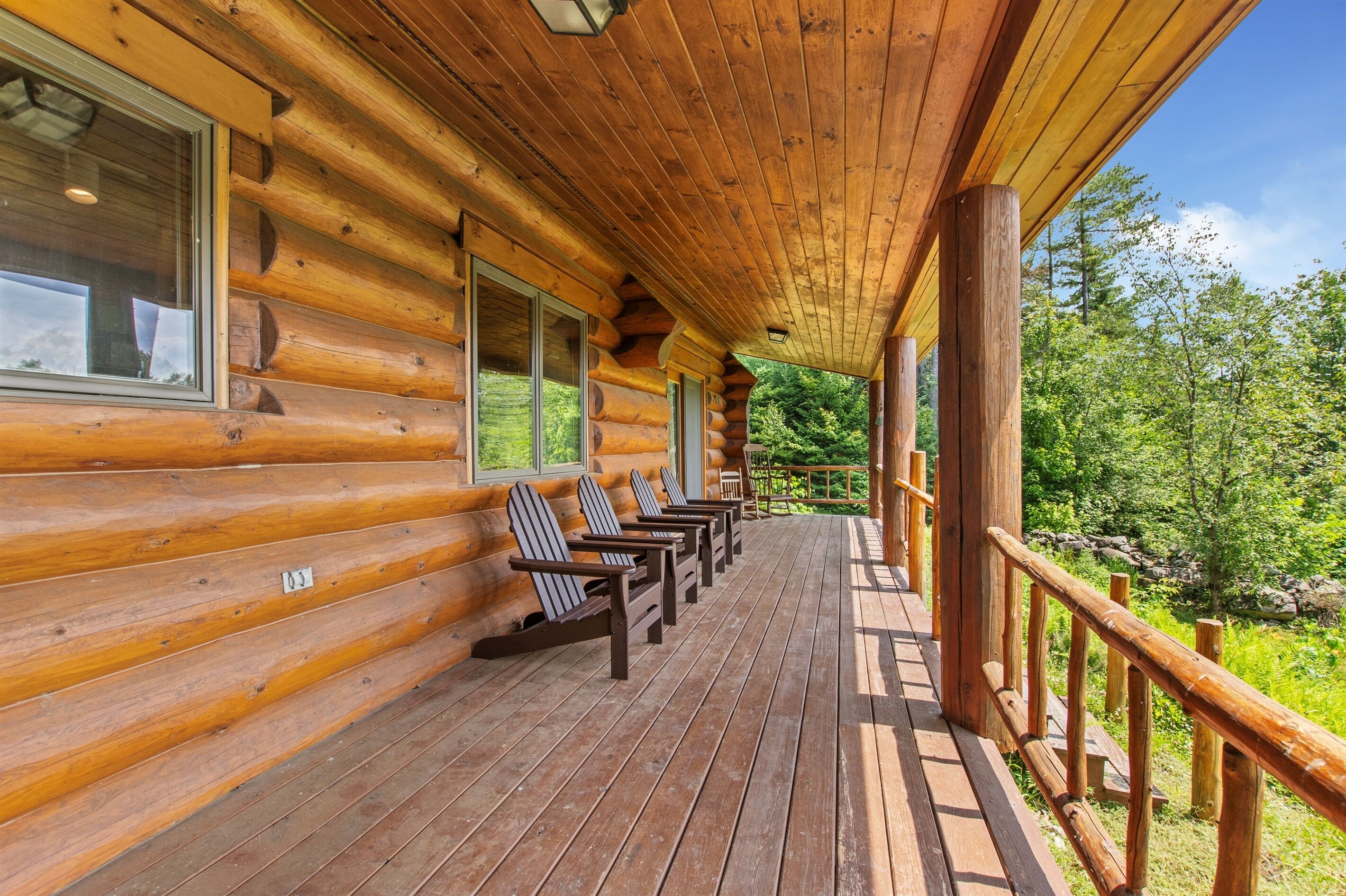 A log cabin with a wooden deck, chairs, and a view of the surrounding forest.
