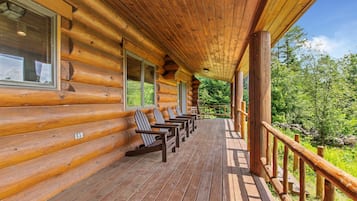 A log cabin with a wooden deck, chairs, and a view of the surrounding forest.