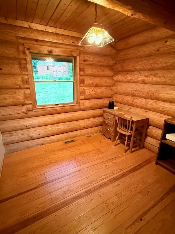 Une chambre dans un chalet en rondins, avec un plancher en bois, une fenêtre donnant sur une structure en bois à l’extérieur, une chaise en bois et une petite commode.