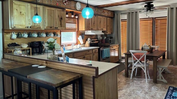 A kitchen with wooden cabinets, a central island, pendant lights, and a dining area with a table and chairs. - Port Clinton Lakeside Escape (Erie Township)