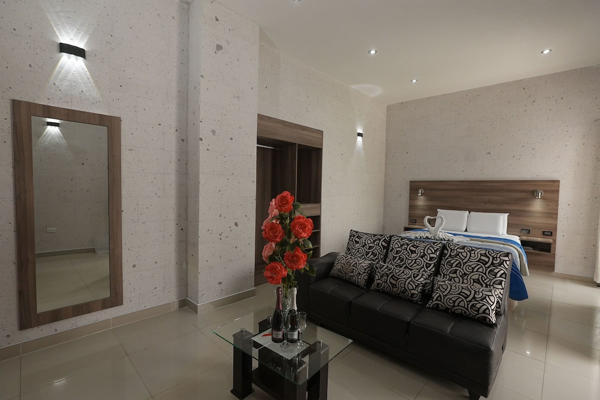 Quarto de hotel moderno com mesa de centro de vidro, vaso com flores, sofá de couro preto, cama com almofadas estampadas e um espelho grande.