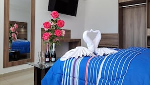 Quarto de hotel com cama, espelho, televisão, um vaso de rosas cor-de-rosa e uma garrafa de champanhe.