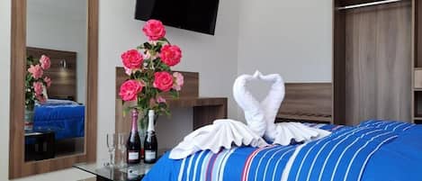 A hotel room with a bed, a mirror, a TV, a vase of pink roses, and a bottle of champagne.