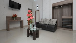 Uma sala de estar moderna com uma mesa de centro de vidro, um sofá preto, uma televisão fixada na parede e um guarda-roupa de madeira.