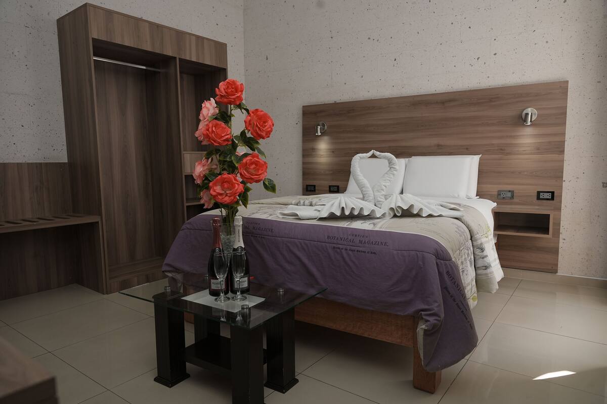 Quarto com cabeceira de madeira, cama com roupa de cama branca, manta roxa e mesa de vidro com um vaso de flores e duas garrafas.