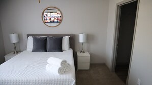 A neatly made bed with white bedding and rolled towels, two bedside tables with lamps, a wall-mounted TV, and a door leading to another room. - 3 mins from Space Center &BridgeStreet. (Huntsville)