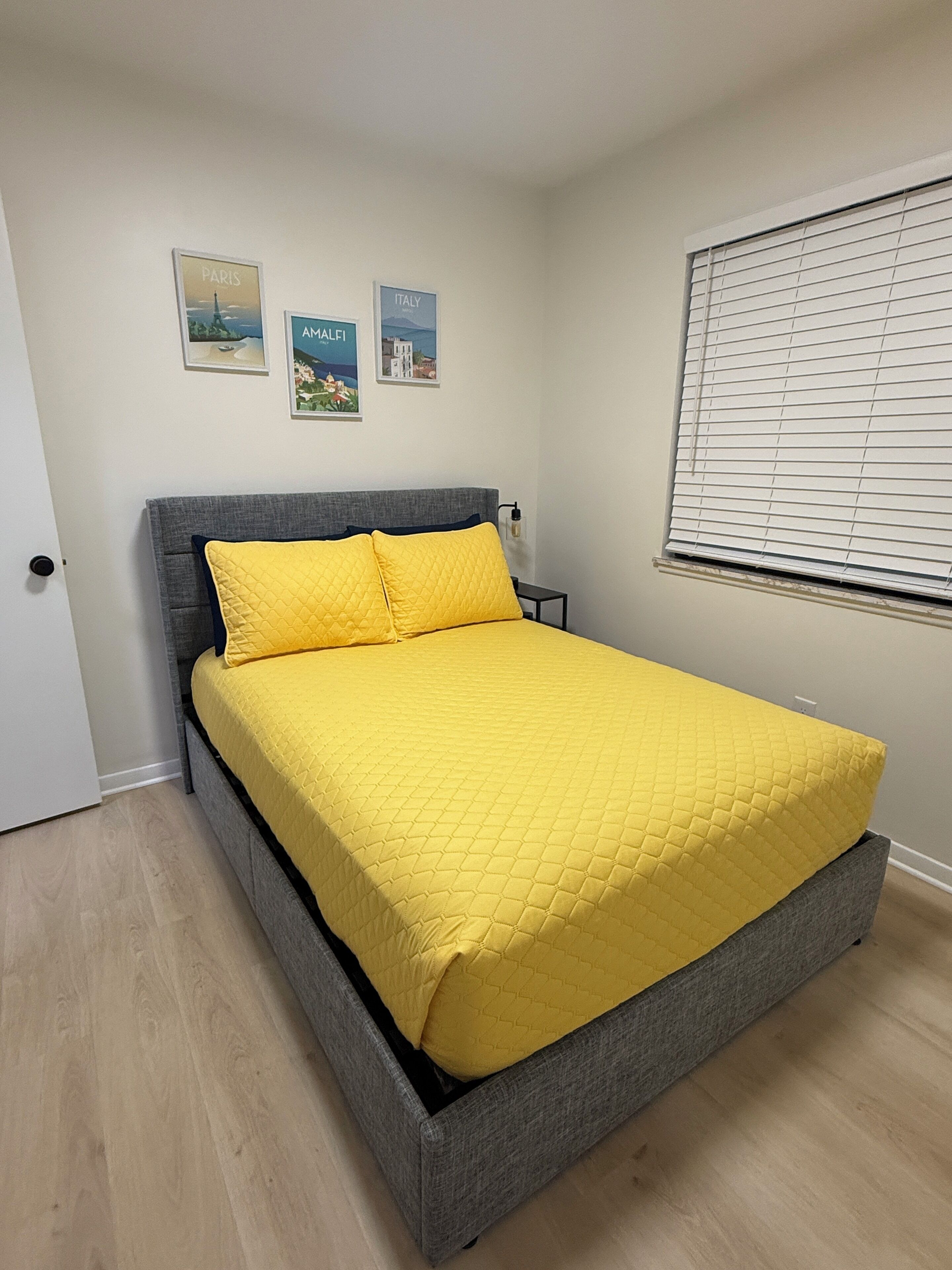 Ein Bett mit einer gesteppten gelben Bettdecke und Kissen, einem grauen Kopfteil und zwei gerahmten Bildern an der Wand.