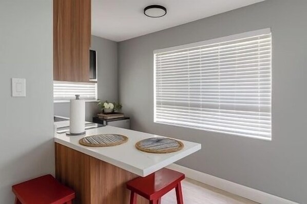 A modern kitchen with a white countertop, two red stools, a wooden cabinet, and a window with blinds.