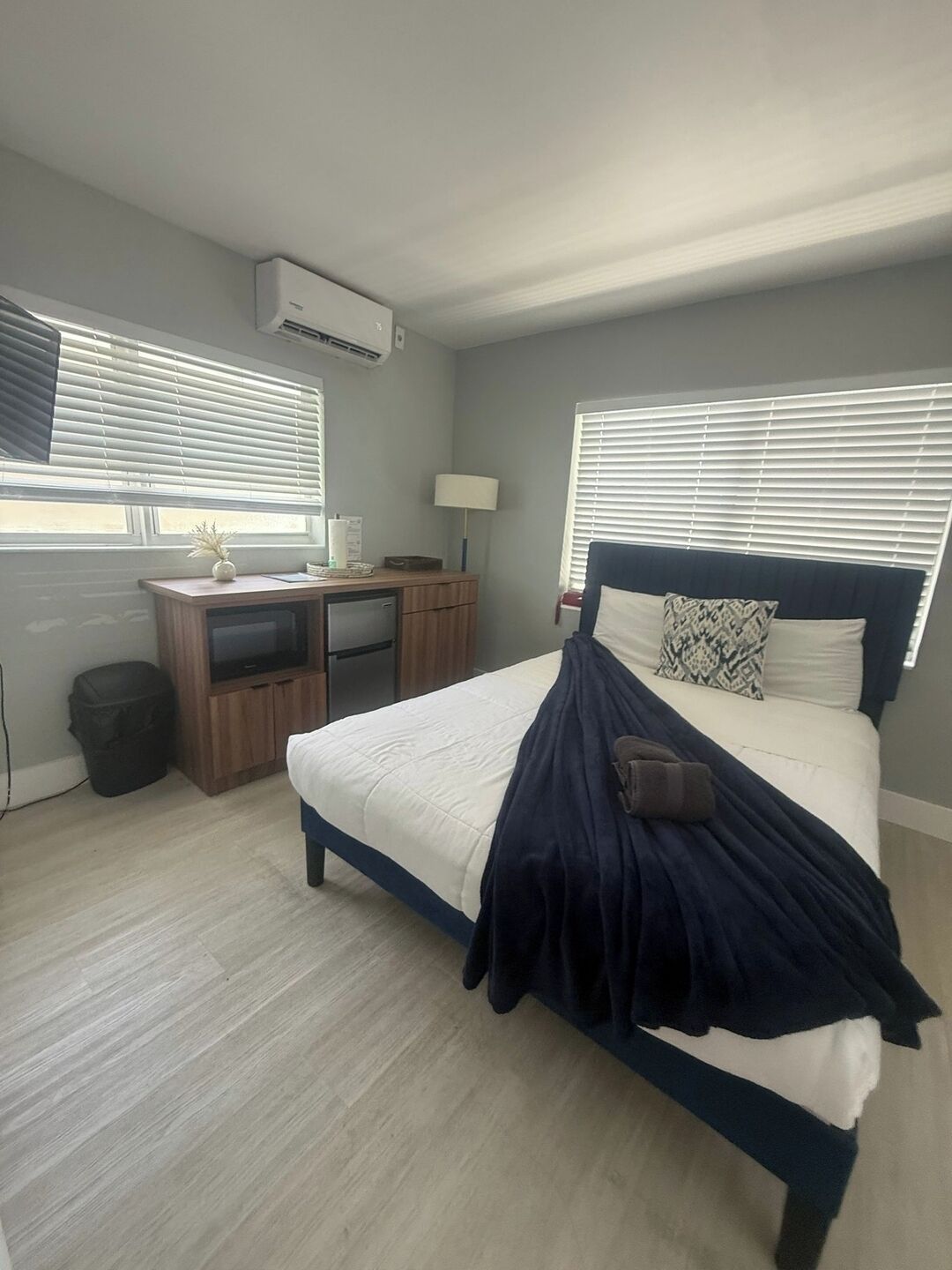 Een slaapkamer met een bed, een bureau en een stoel. Er is een raam met zonwering, een lamp en een airconditioner aan de muur.