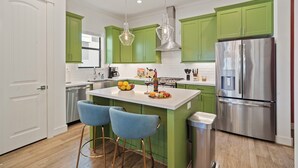 Une cuisine moderne avec des meubles de couleur verte, un réfrigérateur en acier inoxydable et un coin bar avec des chaises bleues.