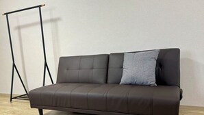 Een donkerbruine leren bank met één lichtgrijs kussen tegen een effen muur en houten vloer.