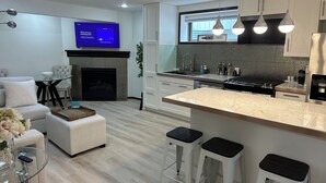 Eine moderne Küche mit Einbauherd, weißen Schränken und einer großen Kochinsel mit Barhocker. An der Wand ist ein Flachbildfernseher angebracht und im Wohnbereich befindet sich ein Kamin.
