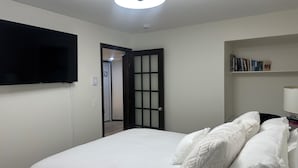 Ein Schlafzimmer mit einem Bett, einem an der Wand befestigten Fernseher, einem Bücherregal und einer Tür zu einem weiteren Raum.
