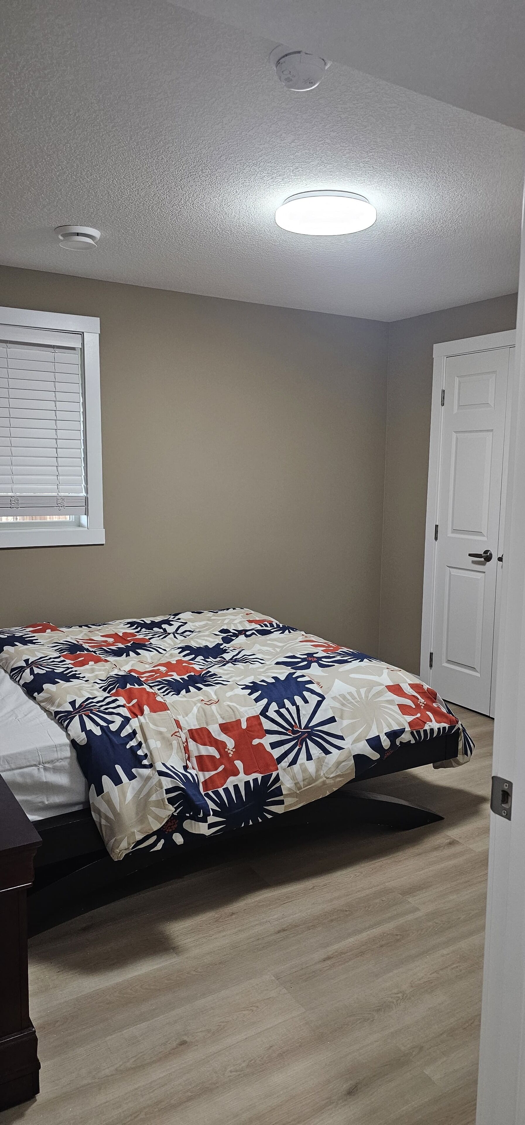 Une chambre à coucher avec un lit, une fenêtre avec des stores, une porte et un luminaire au plafond.