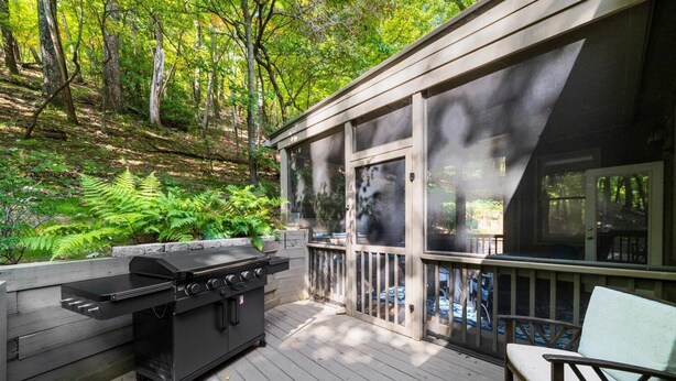 A covered outdoor deck with a grill, a chair, and a view of a forested area.