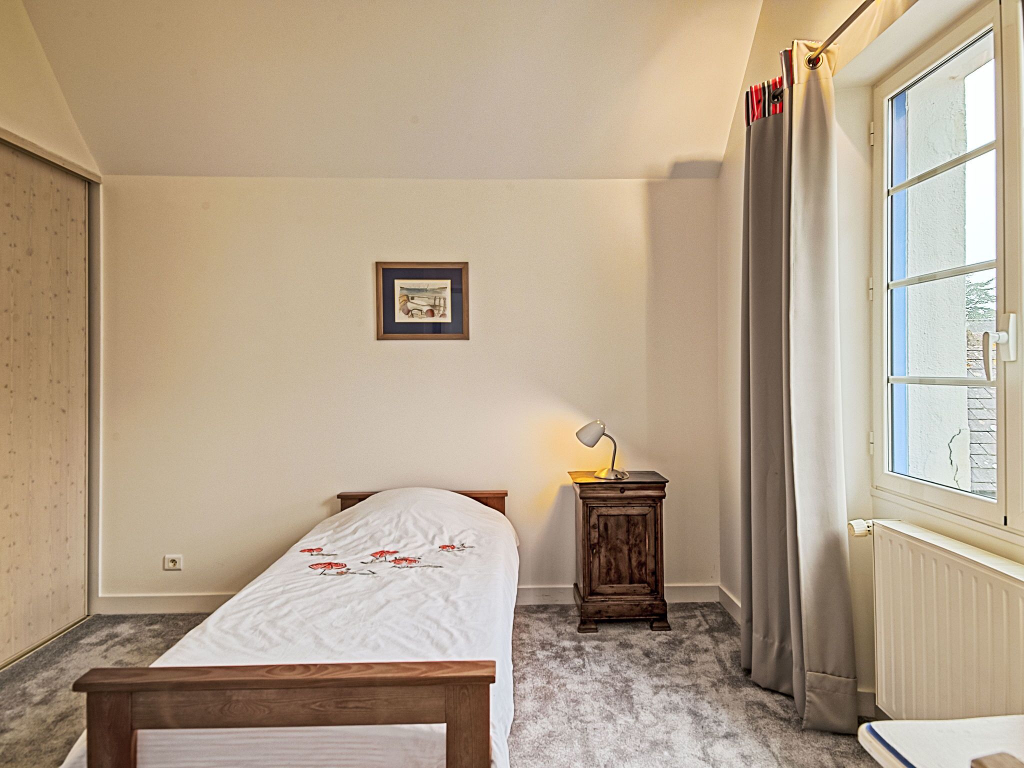 A bedroom with a bed, a bedside table with a lamp, a framed picture on the wall, and a window with curtains.