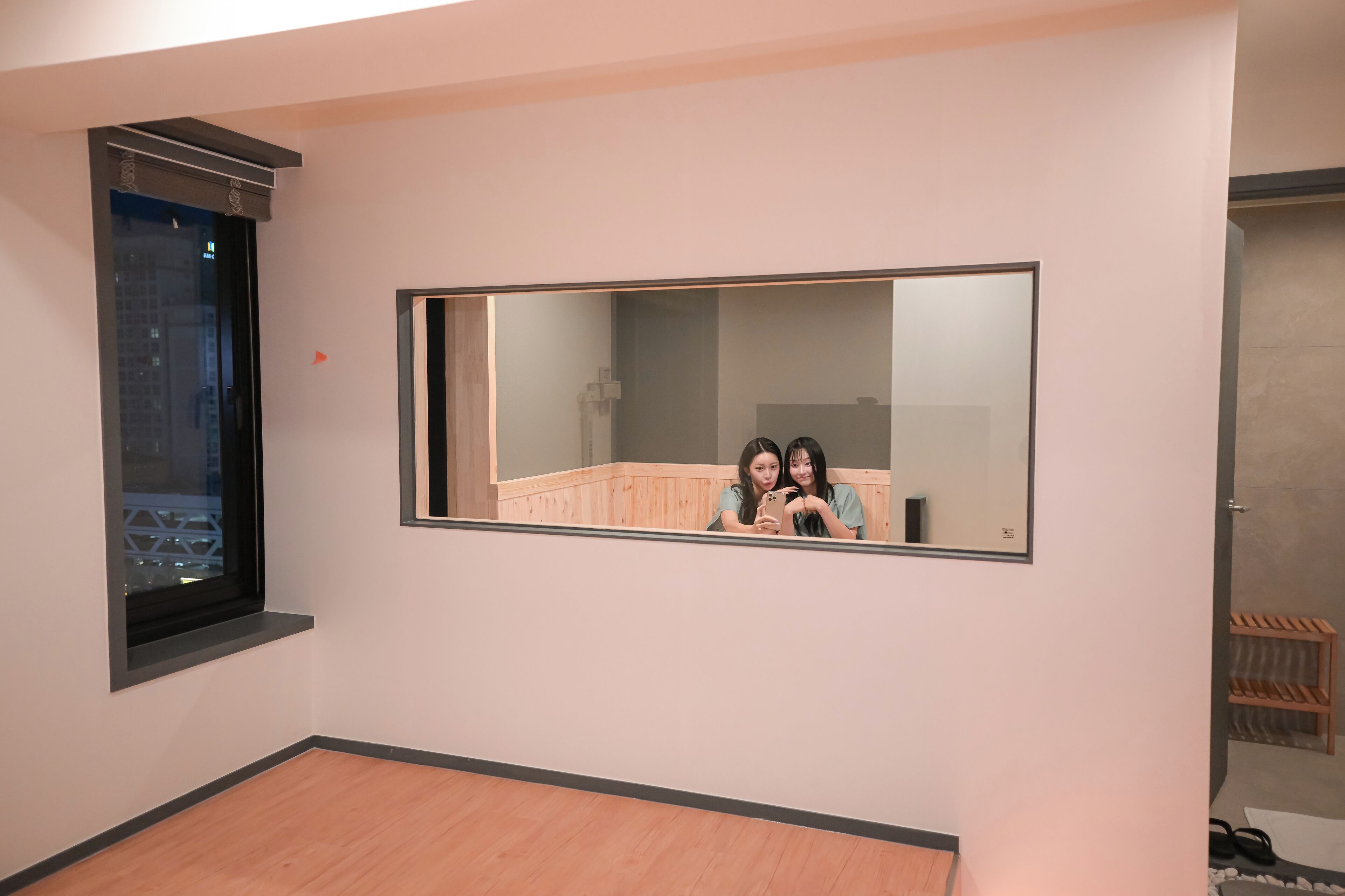 A room with a large mirror on the wall, a window with a black frame, and a wooden floor.