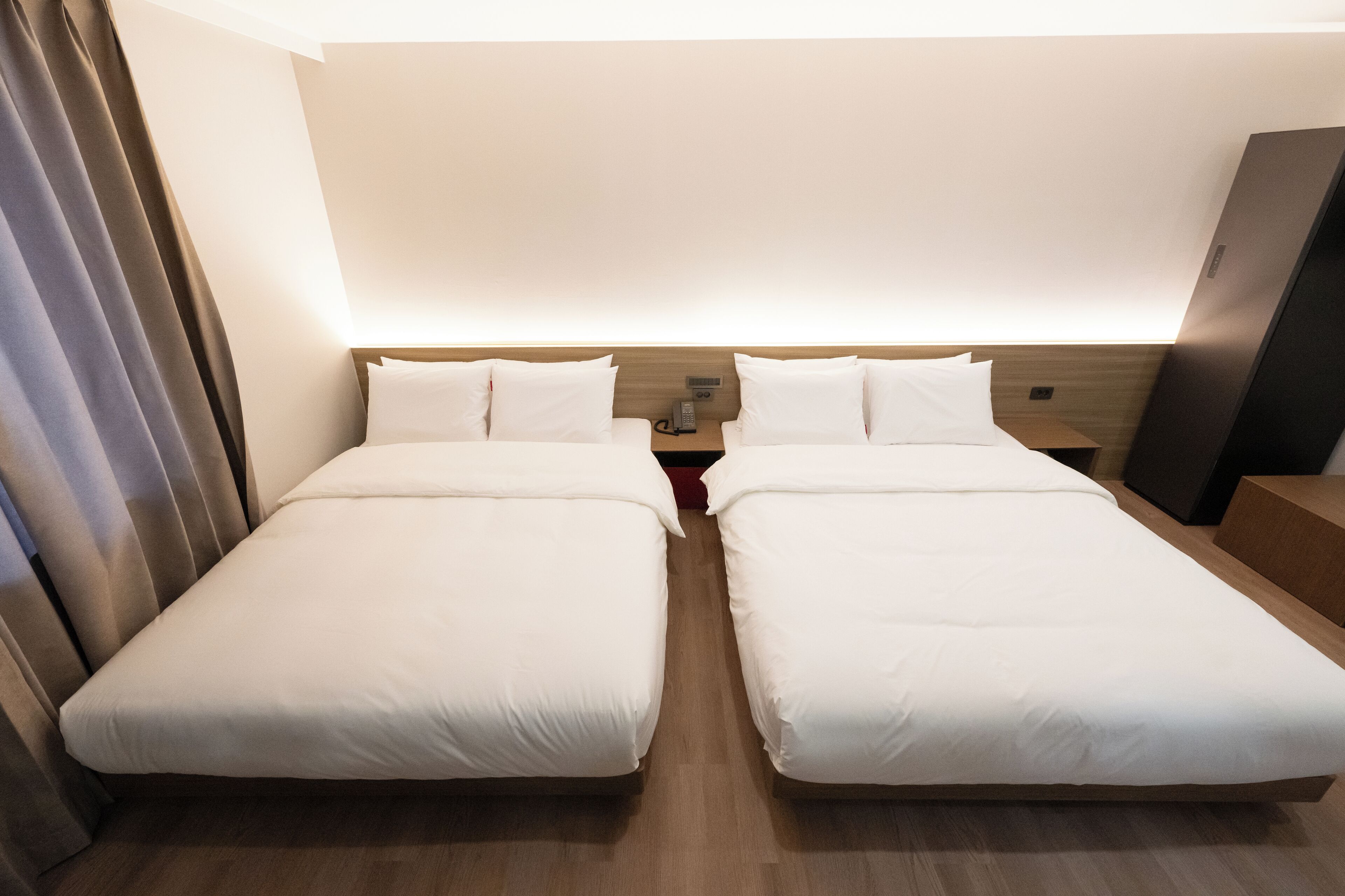 A hotel room with two single beds, a bedside table with a lamp, and a flat-screen TV on the wall.