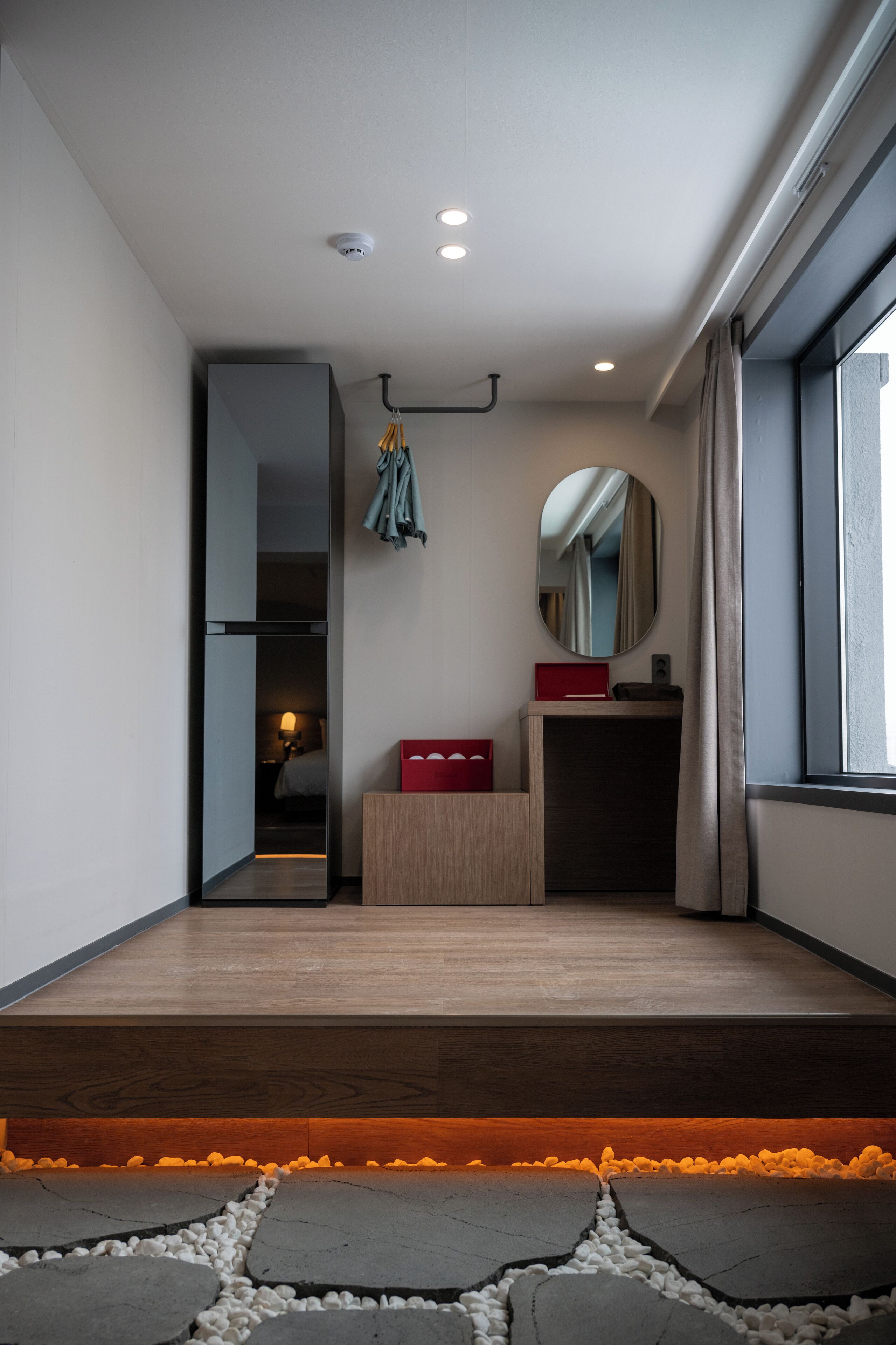 A modern hotel room with a wooden floor, a small wooden desk, a mirror, and a red box.