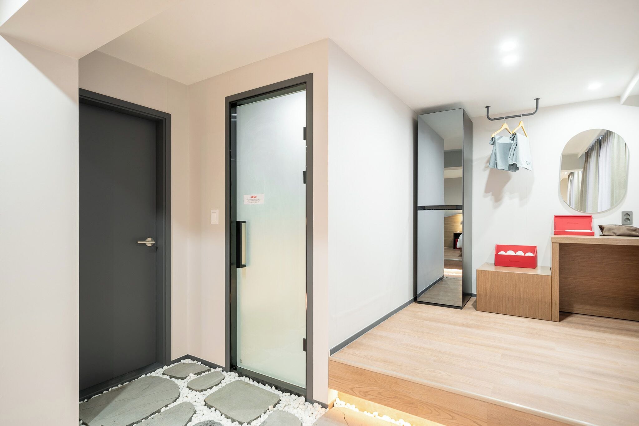Une chambre d’hôtel moderne avec une porte sombre, une porte en verre dépoli, un plancher en bois, un miroir, une coiffeuse en bois et une boîte rouge posée sur une petite table.