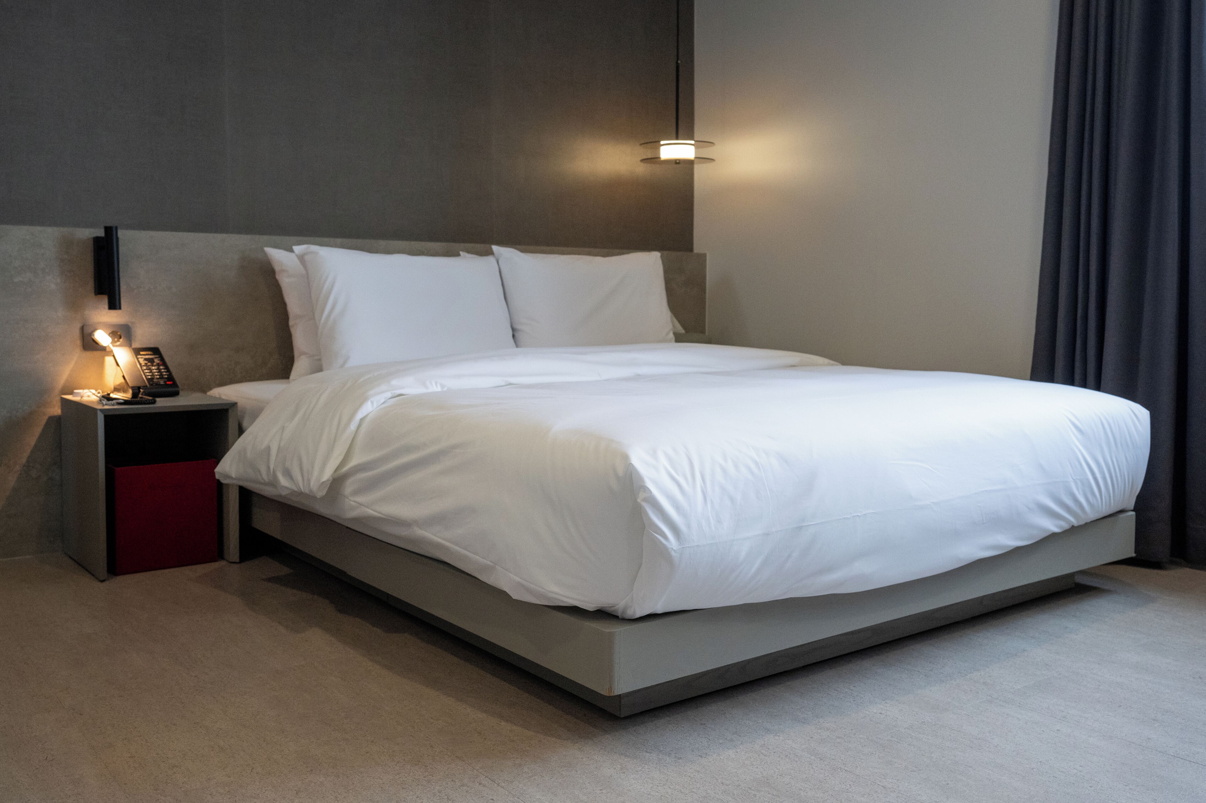 A modern hotel room with a large bed, a bedside table with a lamp, and a wall-mounted light fixture.