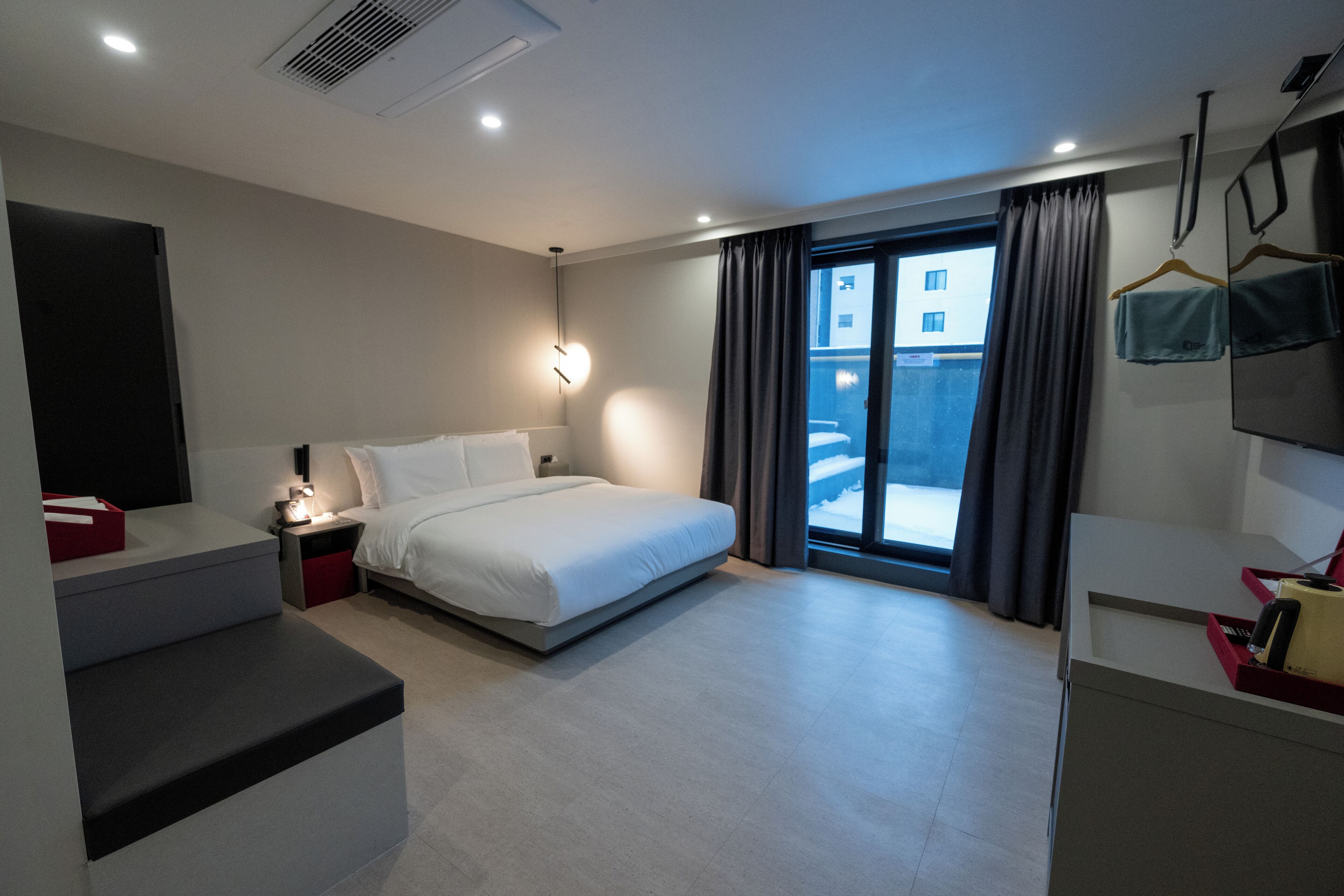 A modern hotel room with a bed, bedside table, and a view of the outside.