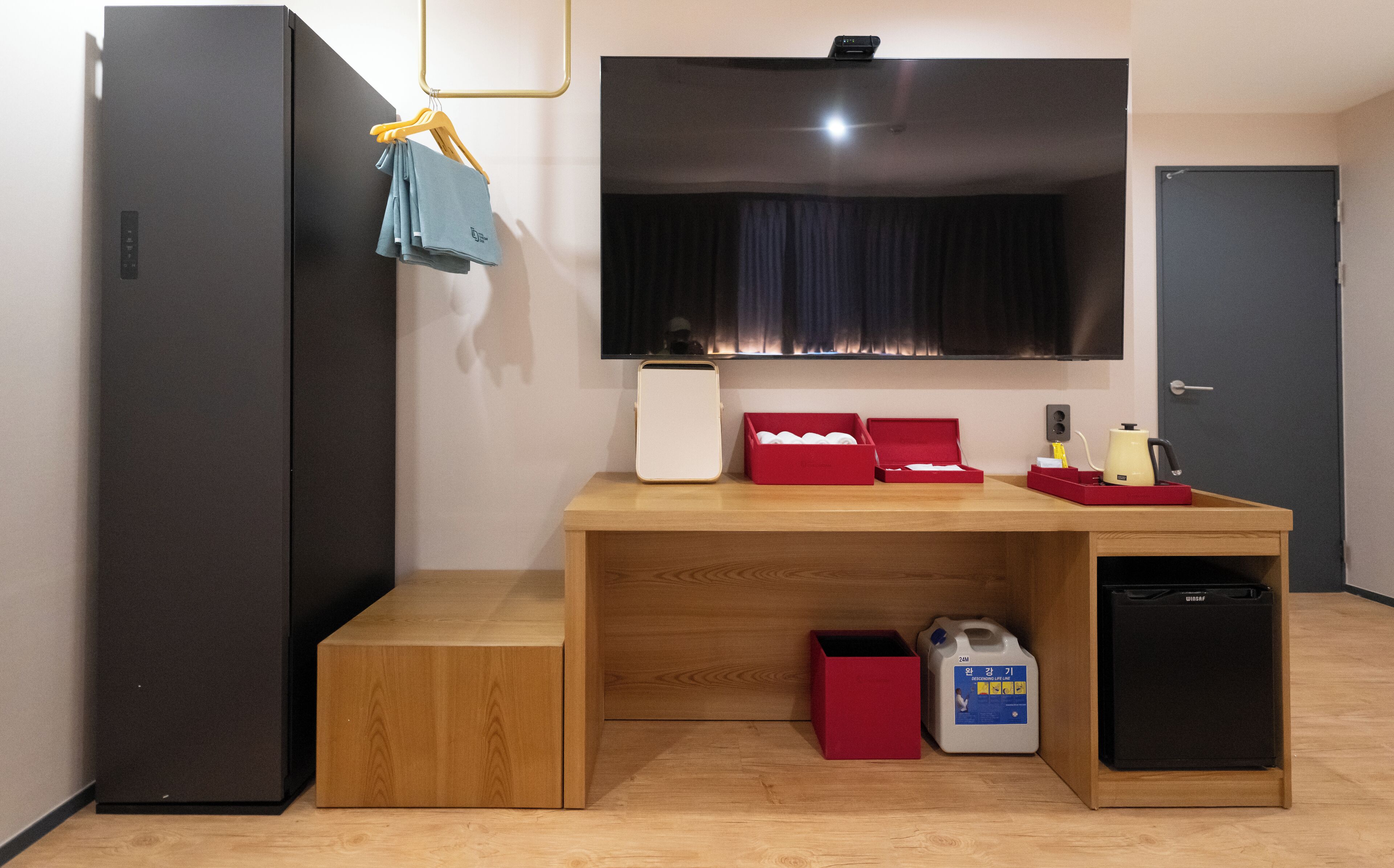 Ein kleines Hotelzimmer mit einem Holztisch, einem Flachbildfernseher, einer Minikühlschrank, einer roten Schachtel und einer Milchtülle.
