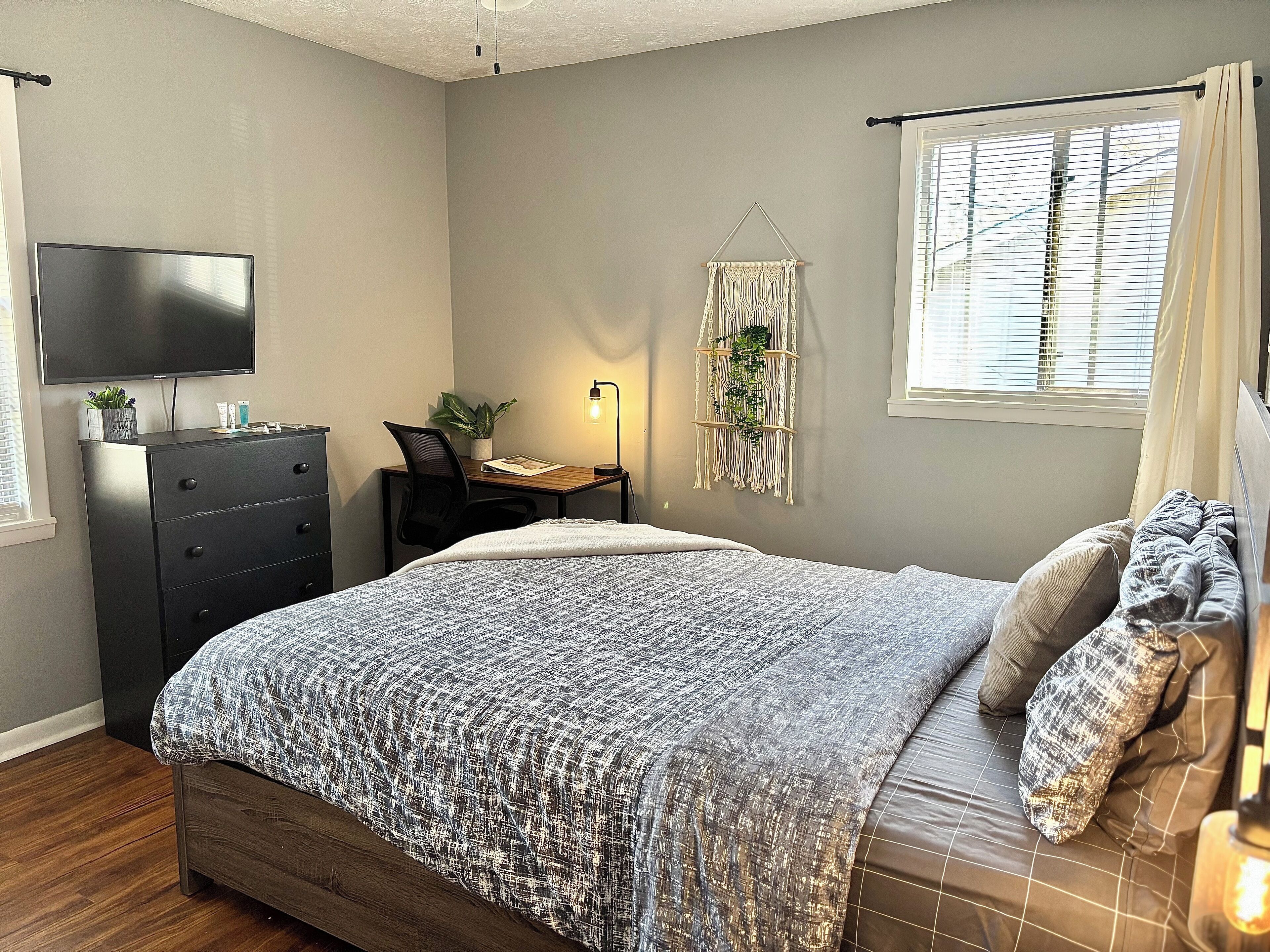 Quarto com cama, escrivaninha, cadeira, cômoda, televisão, janela com persianas e planta na parede.