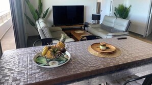 Un comedor con una mesa dispuesta para dos personas, que incluye una fuente de frutas y un plato pequeño con salsas. En el fondo, un televisor de pantalla plana está montado en la pared, y en la zona de estar contigua hay un sofá y una silla.
