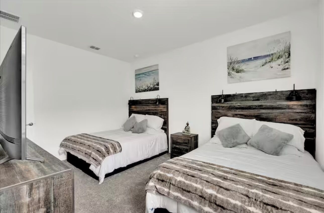 Quarto com duas camas, cabeceira de madeira, mesa de cabeceira, televisão e obras de arte nas paredes.
