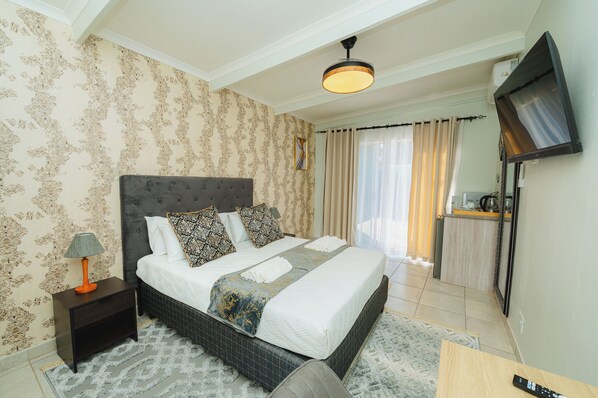 A neatly arranged bedroom with a large bed, a bedside table with a lamp, a TV mounted on the wall, and a small kitchenette area. - Mawo Luxury Boutique (Johannesburg)