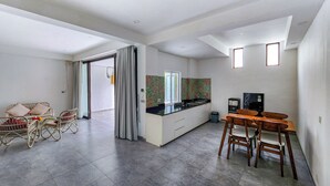 Una cocina moderna con encimera negra, armarios blancos y un ventanal pequeño. Hay una zona de comedor con mesa y sillas de madera, y un sofá de mimbre con cojines.