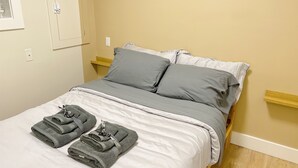 Een net opgemaakt bed met grijze beddengoed en kussens, een houten hoofdbord en twee opgevouwen handdoeken op het bed.