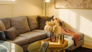 Een gezellige woonkamer met een hoekbank, een vloerlamp, een bijzettafeltje met decoratieve objecten en een groot abstract schilderij aan de muur.