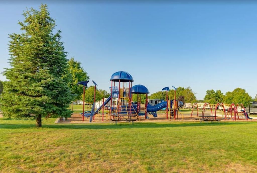 A playground with slides and climbing structures, surrounded by grass and trees.
