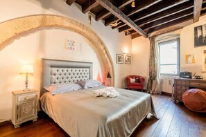 Un dormitorio con cabecera capitonada, cama con toallas enrolladas, mesita de noche con lámpara, una silla roja y un aparador.