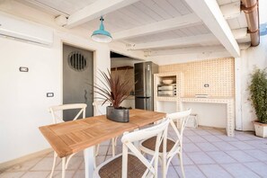 Un comedor con mesa de madera y sillas blancas, una planta en maceta y una cocina con salpicadero decorativo.
