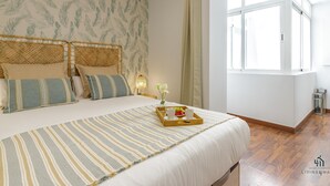 A bedroom with a large bed, a window with curtains, a bedside table with a lamp, and a tray with fruit and cups.