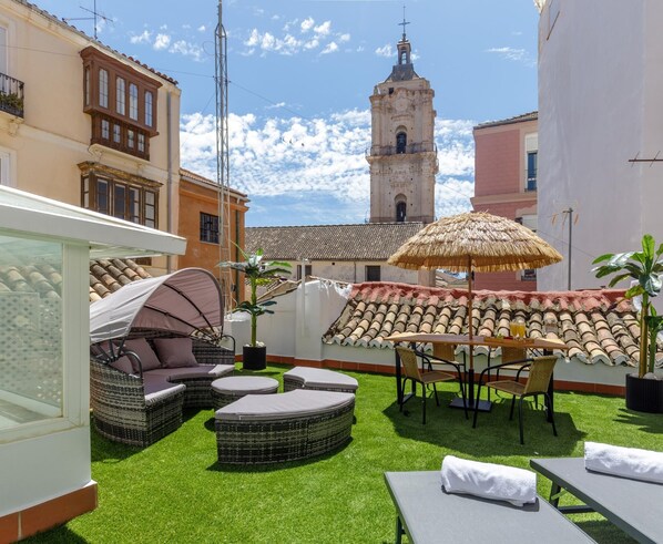 Una terraza en la azotea con mobiliario exterior, un paraguas de paja y vistas a un edificio histórico con una torre de campanas.