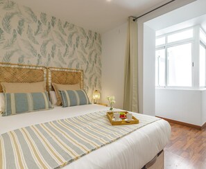 Un dormitorio con una cama grande, una ventana con cortinas, una mesita de noche con lámpara y una bandeja con frutas y tazas.