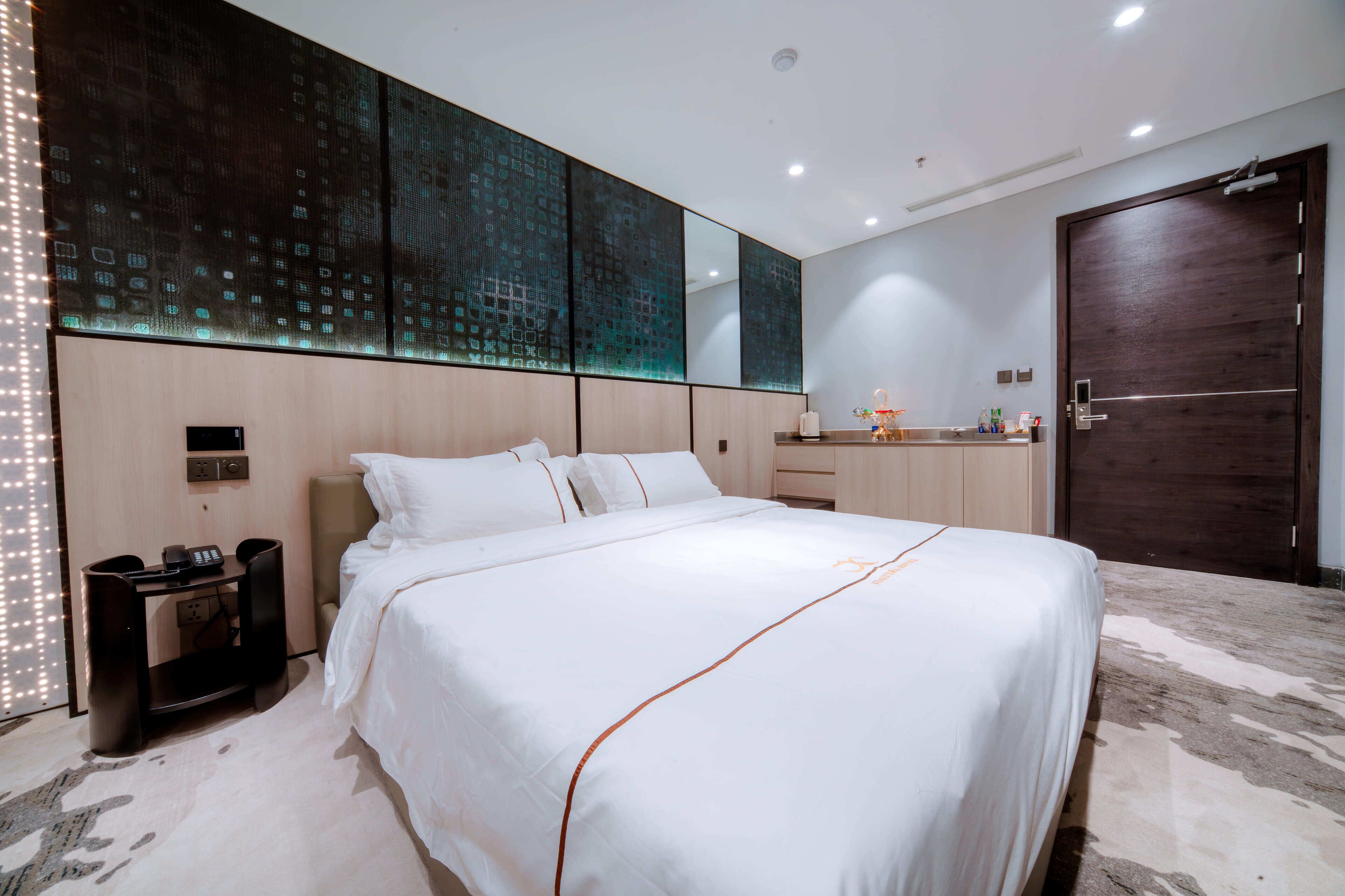 A modern hotel room with a large bed, a desk with a phone, a mirror, and a wooden door.