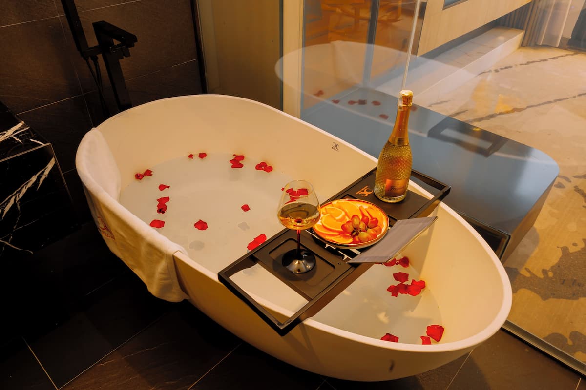A bathtub with rose petals, a bottle of champagne, and a glass of wine.