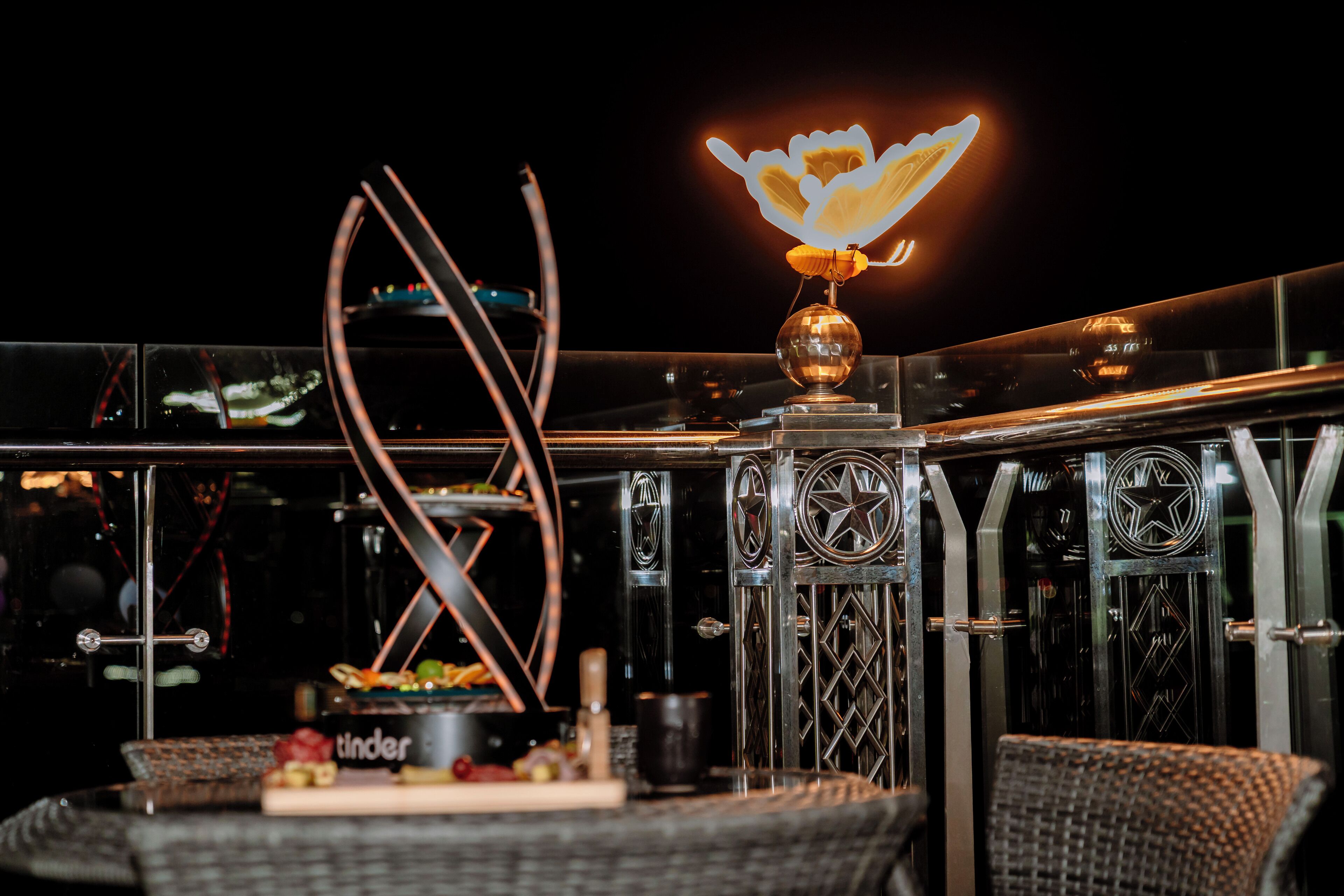 A modern outdoor seating area with a decorative metal sculpture, a table with a cake, and a neon flower sign.
