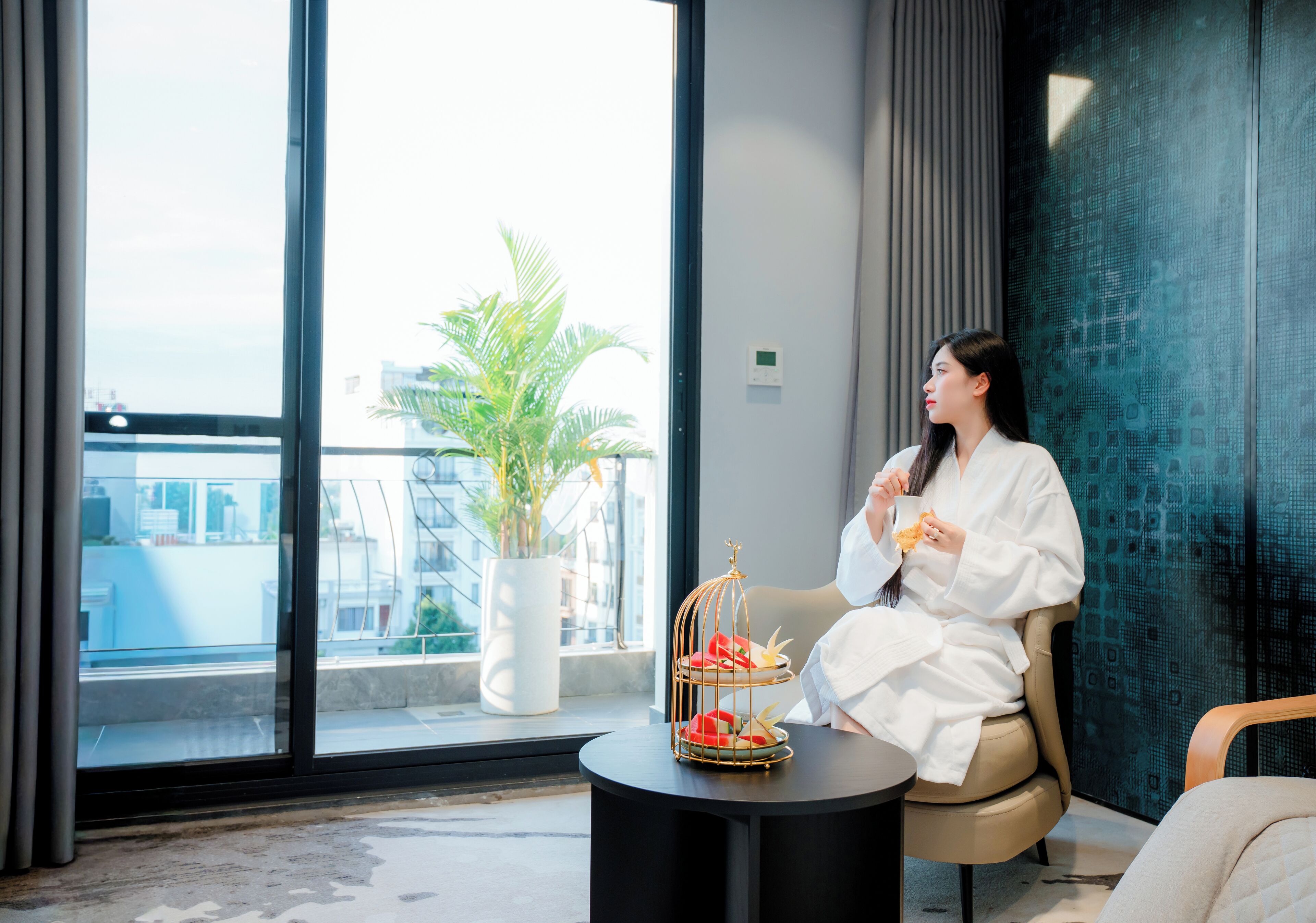 A woman in a white robe sitting by a window with a view of a cityscape, a plant, and a tray with food.