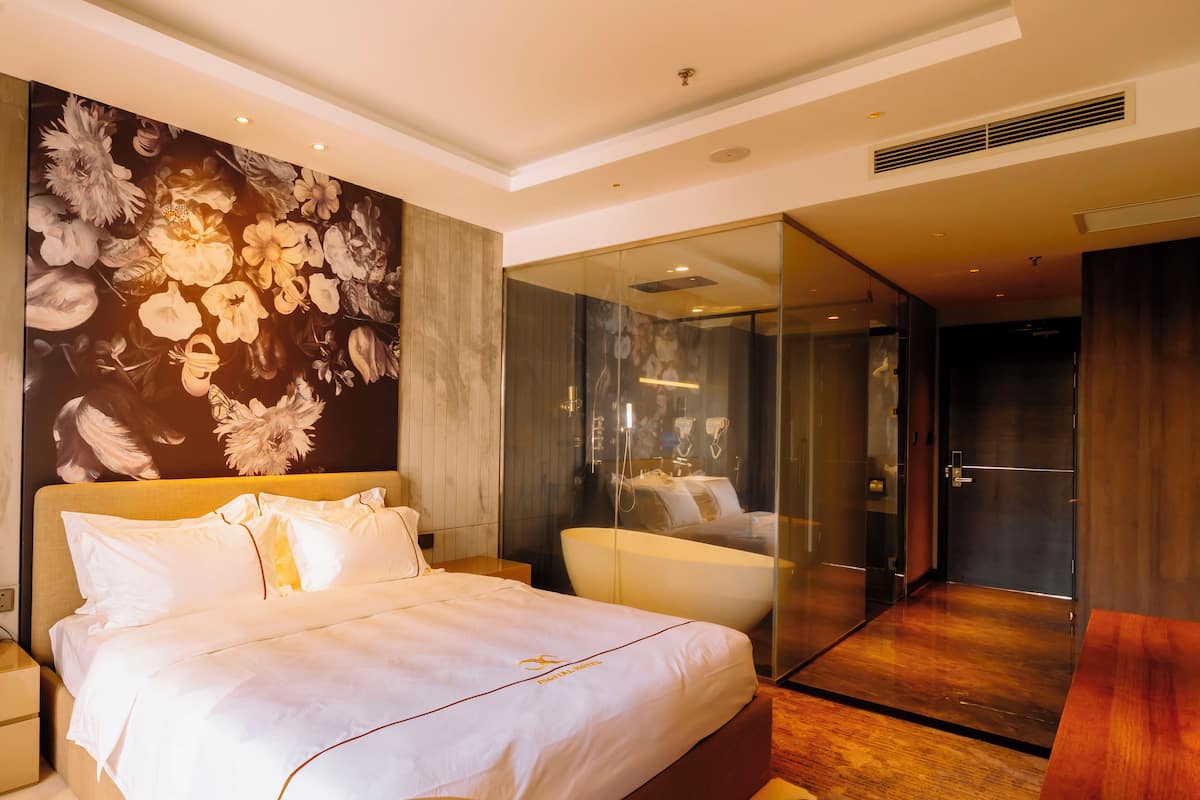 A modern hotel room with a large bed, a glass-enclosed bathroom, and a floral mural on the wall.