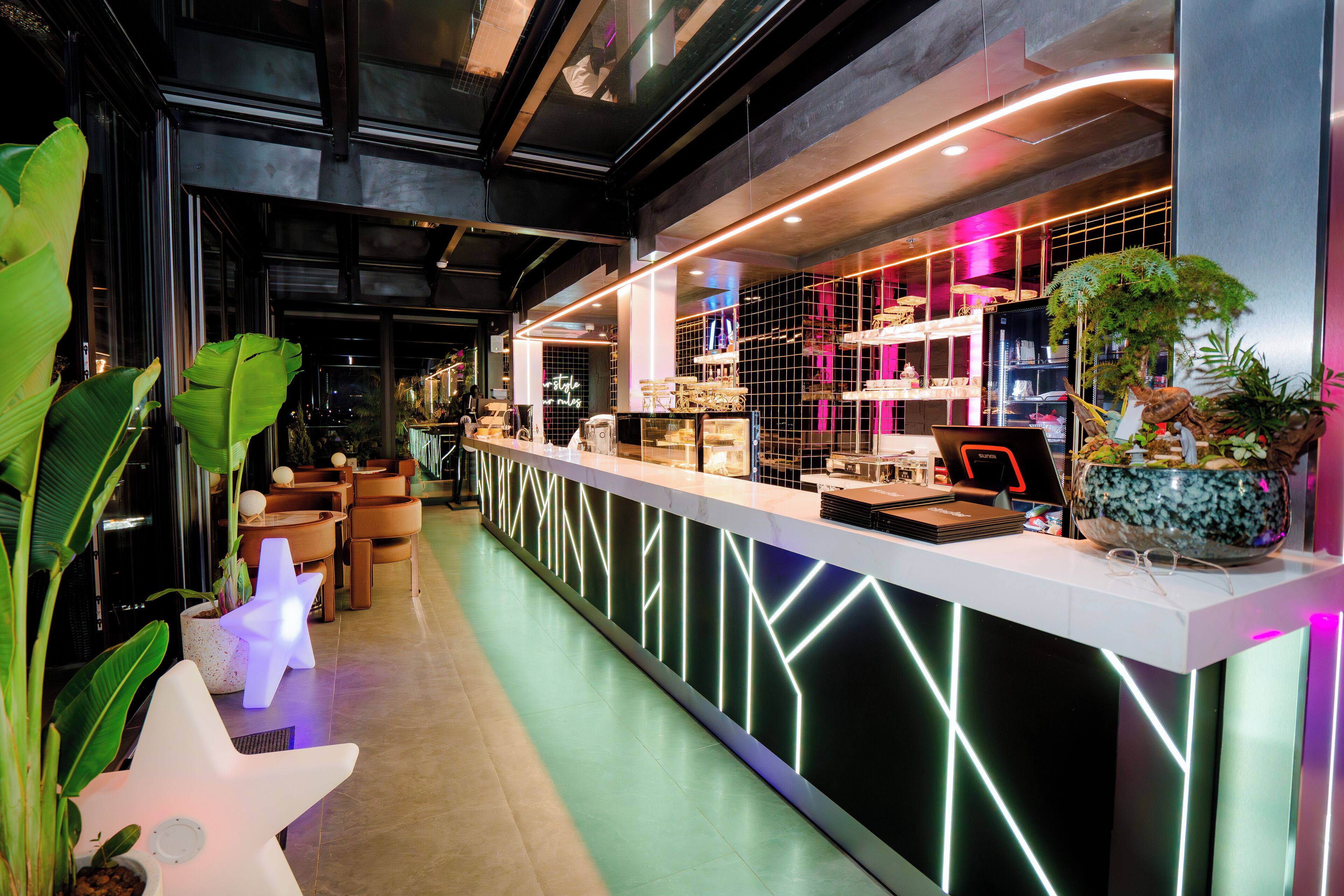 A modern bar area with a neon-lit counter, seating, and potted plants.