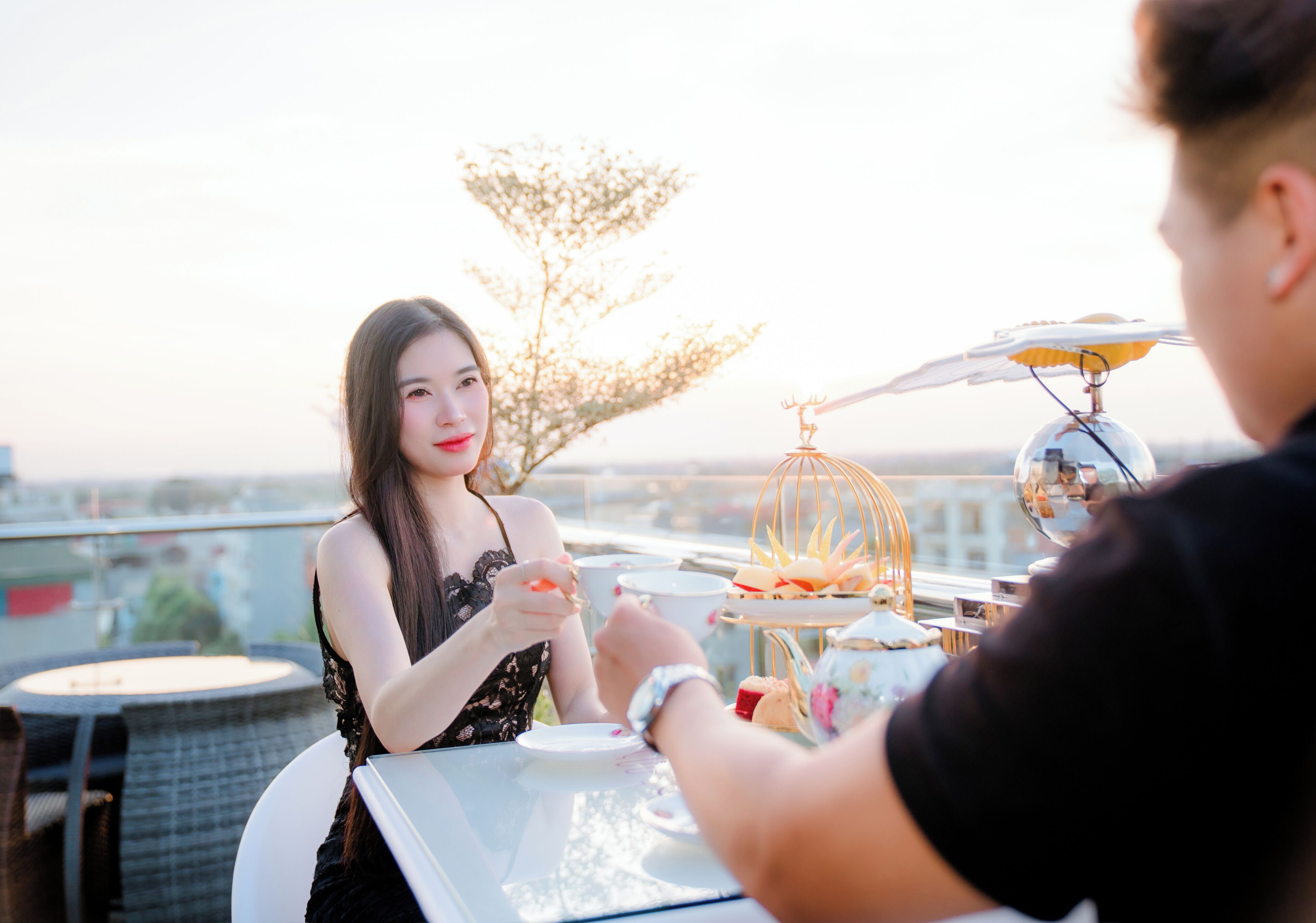 A woman seated at a table with a tea set, a decorative birdcage, and a city view in the background.