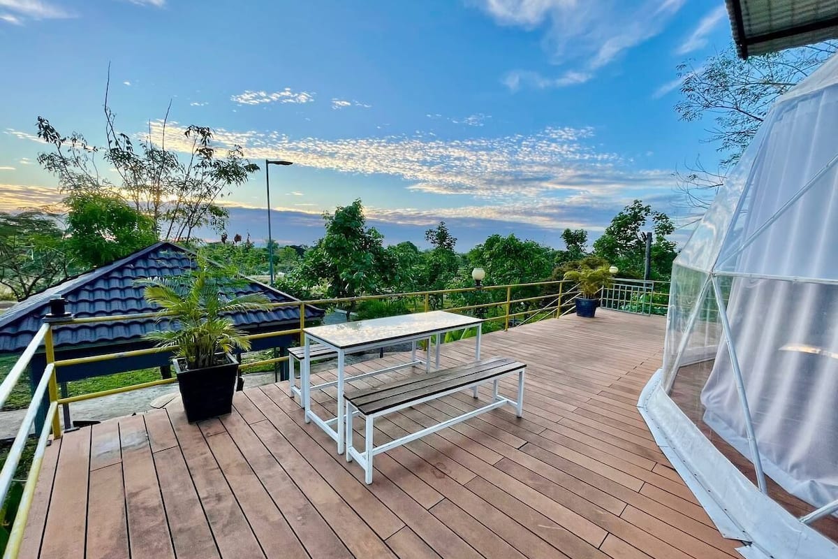 Une terrasse en bois avec une table et un banc, entourée de verdure et sous un ciel dégagé.