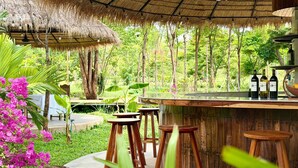 Een terras met houten krukken, een rieten dak en uitzicht op weelderige begroeiing.